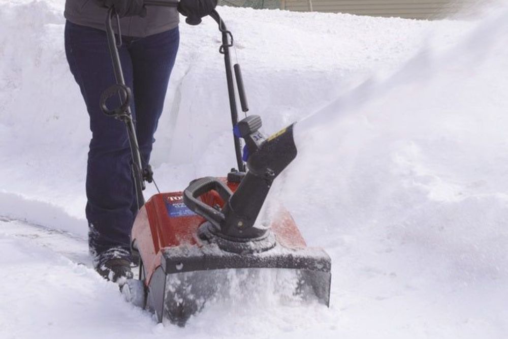 A woman operates a snow blower, clearing a path through freshly fallen snow in a residential area.